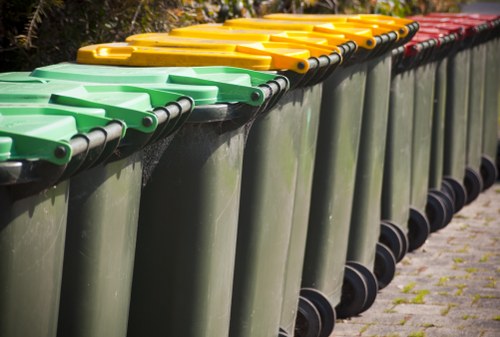 A row of large industrial waste bins aligned along a paved pathway, with the majority of the bins in dark green color. The bins have flat, smooth tops made of durable plastic, each fitted with a matching lid that is either yellow or red, indicating different waste categories. The yellow-lidded bins are positioned towards the front, followed by the red-lidded ones further along the line. The bins are slightly tapered towards the bottom and mounted on black caster wheels for mobility. The background features a blurred outdoor environment, possibly in a commercial or residential area, with some greenery visible. The scene appears to be in daylight, and the bins are lined neatly, reflecting a well-maintained waste disposal area, typical in towns like Tunbridge Wells where efficient rubbish management is essential. This image relates to rubbish removal services such as those offered by Commercial Waste Tunbridge Wells, highlighting the importance of organized and professional waste collection and disposal across areas like TN postcode regions.