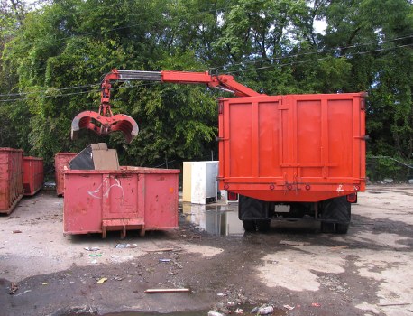 The image shows a waste collection scene with a large red skip bin positioned on a gravel surface, partially behind a red waste collection truck parked to its right. The skip bin appears to be made of metal with a weathered, painted finish and is filled with mixed rubbish, some of which is visible slightly protruding from the top. An industrial robotic arm, painted in red and positioned above the skip, is equipped with a mechanical grabber or claw, which is actively engaged in lifting or emptying the contents of the skip. Surrounding the scene, there are a few more red waste bins or skips in the background, set against a backdrop of dense green trees and foliage, indicating an outdoor environment. The ground is uneven with some scattered debris and what appears to be wet patches or stains, typical of a site where waste collection or disposal takes place. The overall setting is outdoors, likely within a commercial or industrial waste management area, consistent with services provided by Commercial Waste Tunbridge Wells in the local area.