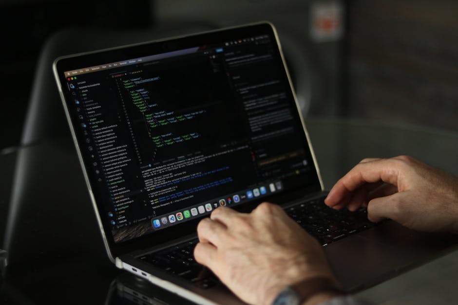 A person's hands are shown typing on a closed laptop placed on a dark surface. The laptop screen displays lines of code with syntax highlighting in various colors, including green, blue, and white, ag